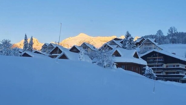 Eine Reihe von Holzhütten unter einer Schneedecke, im Hintergrund ein Berg.