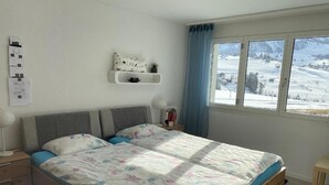 Ein Schlafzimmer mit einem Bett, einem Nachttisch, einer Lampe, einem Fenster mit Blick auf eine verschneite Landschaft und einem Wandregal mit einem dekorativen Gegenstand.