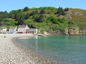 Ein Hotel an der Küste mit einem Kiesstrand, einem Steg und einem bewaldeten Hügel.
