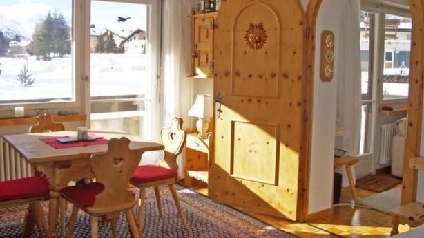 A cozy room with a wooden cabinet, a dining table with chairs, and a view of a snowy landscape through large windows.