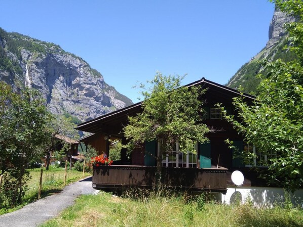 A chalet-style building with a balcony, surrounded by lush greenery and mountains in the background.