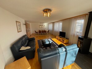 Ein modernes Wohnzimmer mit einem schwarzen Ledersofa, einem Holztisch und einem Fernseher. Im Hintergrund ist eine Essecke mit Tisch und Stühlen zu sehen.