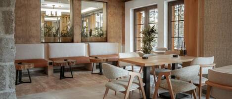 A modern dining area with wooden tables and chairs, large windows, and a stone wall.