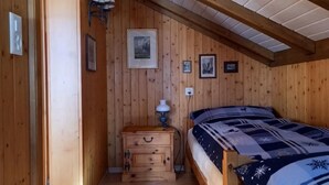 Et hyggeligt rum med trævægge, en seng med et mønstret dynebetræk, et natbord med en lampe og billeder i rammer på væggen.