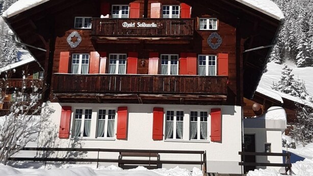 En traditionel chalet-stil bygning med træfacade og røde skodder, med teksten "Chalet Soldanella".