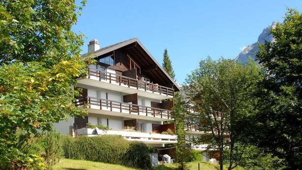 Un edificio a più piani con balconi, immerso nel verde con una montagna sullo sfondo.