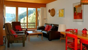 Un accogliente soggiorno con divano, poltrona e tavolino, una zona pranzo con tavolo e sedie, e ampie finestre con vista sulle montagne.