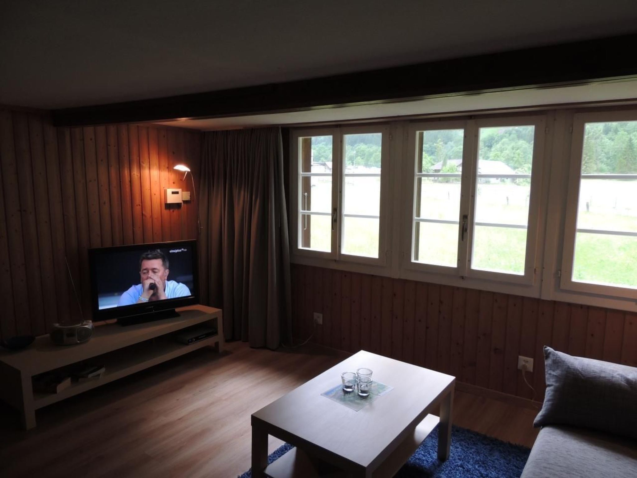 Una habitación con una televisión sobre un soporte de madera, una mesita con vasos y una ventana con vistas a zonas verdes.