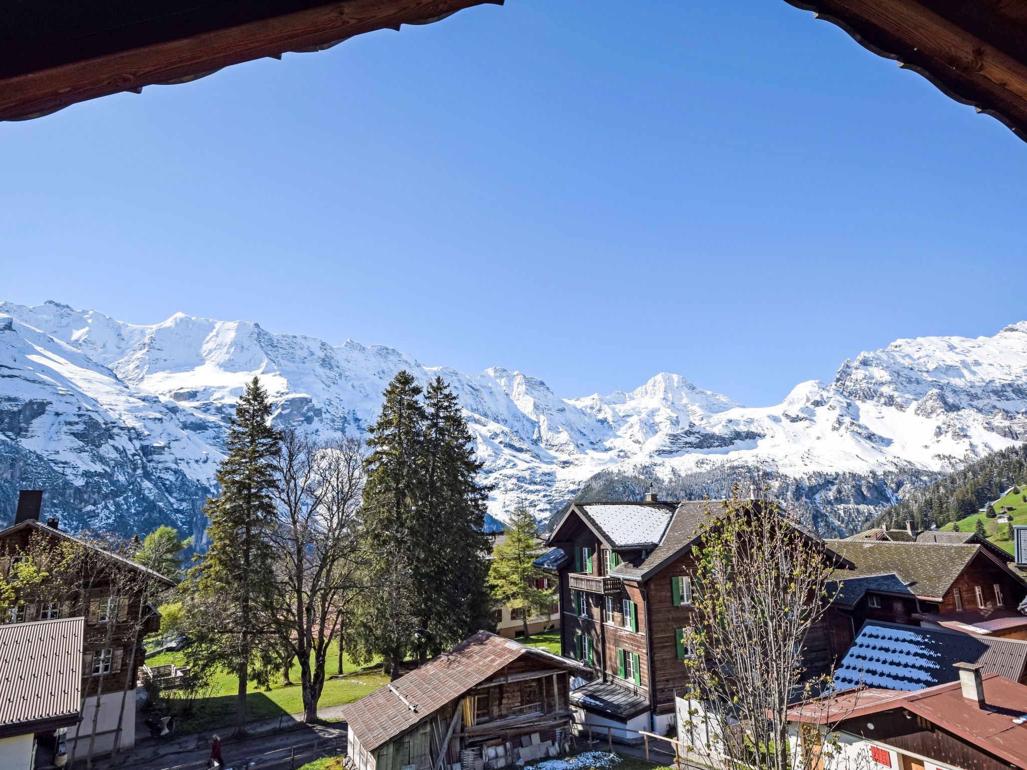A mountain village with traditional wooden houses, a clear blue sky, and snow-covered peaks in the background.