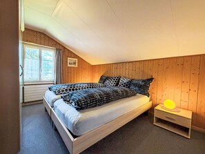 Een slaapkamer met een houten schuine plafond, een bed met beddengoed met een patroon, een nachtkastje met een lamp en een raam met gordijnen.