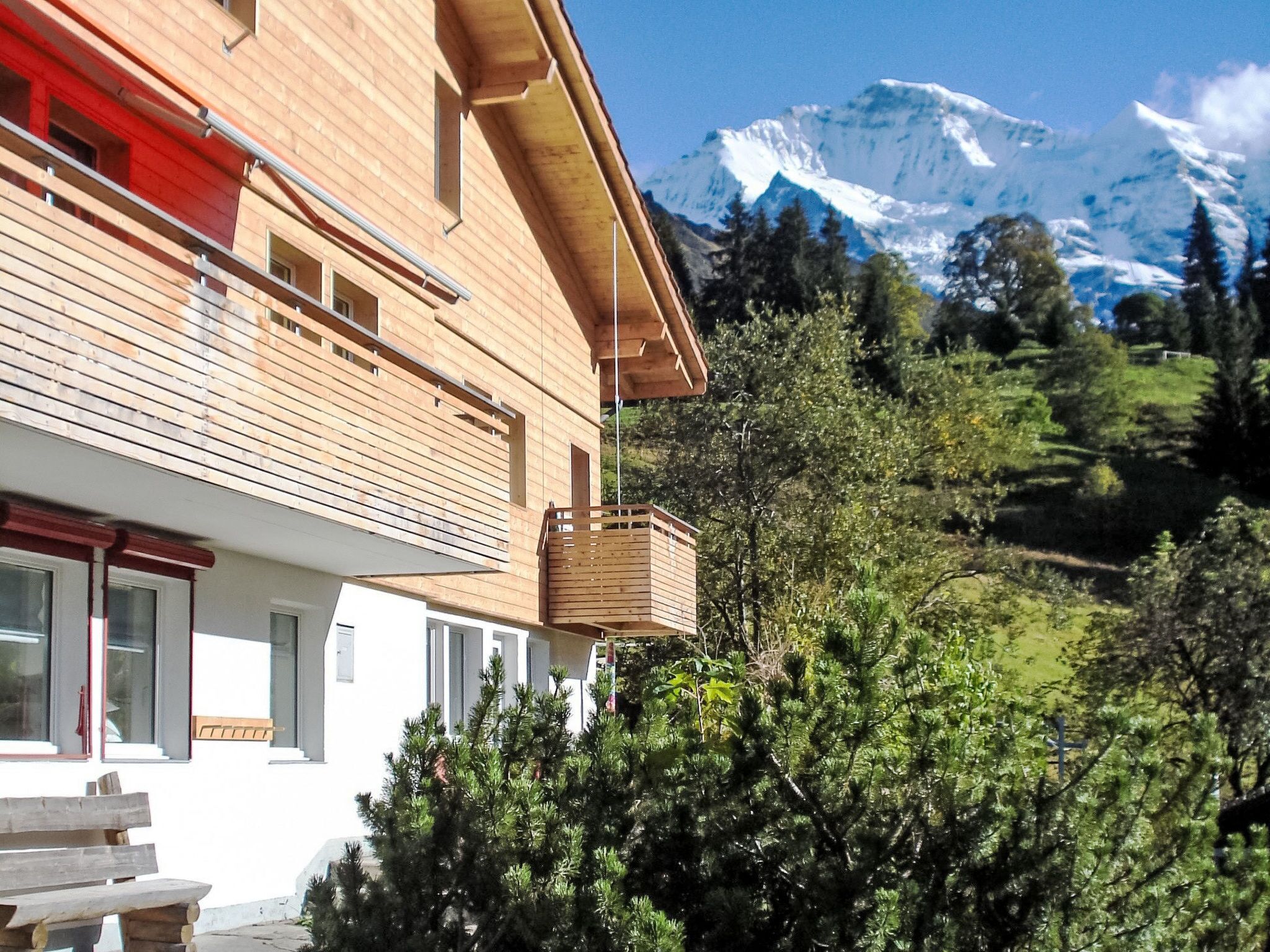 A modern wooden building with a balcony, surrounded by lush greenery and a mountain in the background.