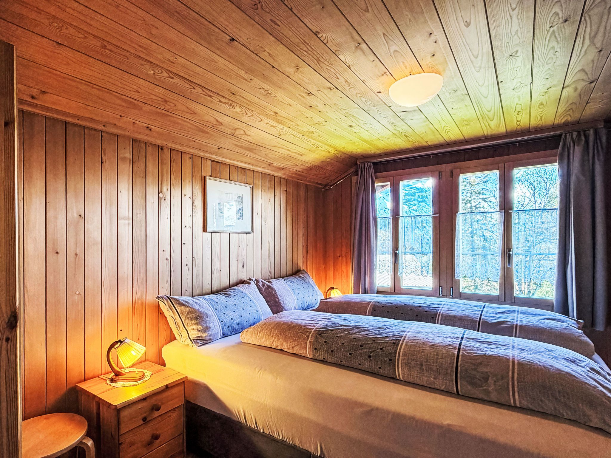 Habitació amb panells de fusta, un llit, una tauleta de nit, una làmpada i una finestra amb cortines.