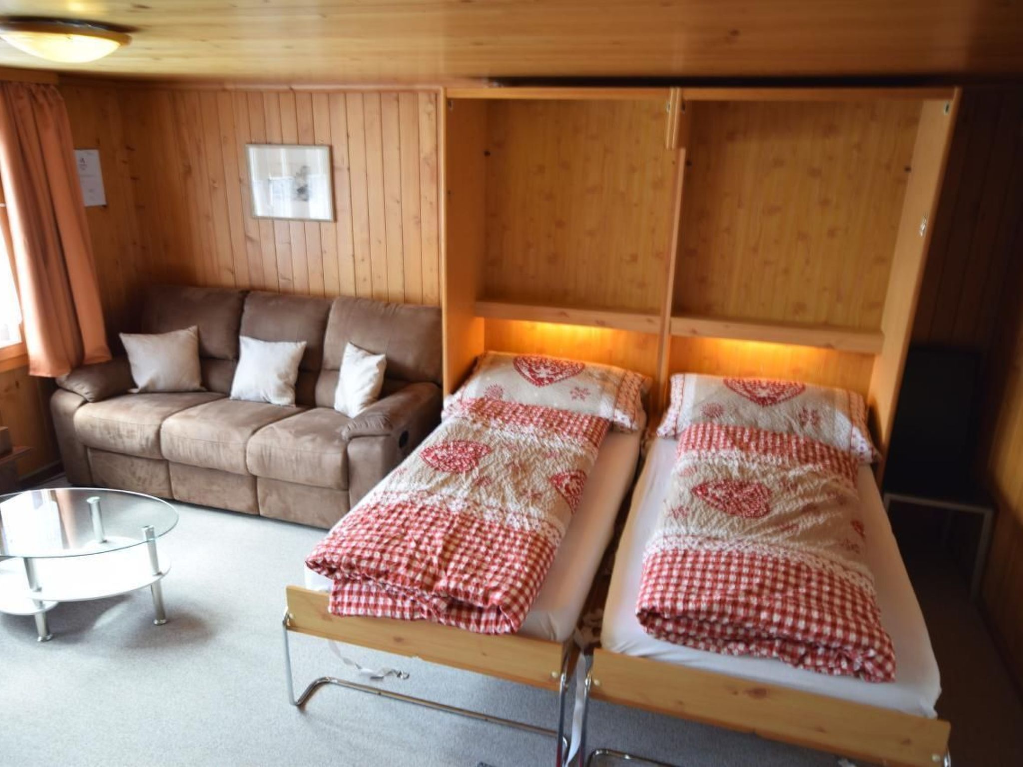 A cozy room with a wooden interior, featuring a sofa, two single beds with red and white checkered blankets, and a small table.