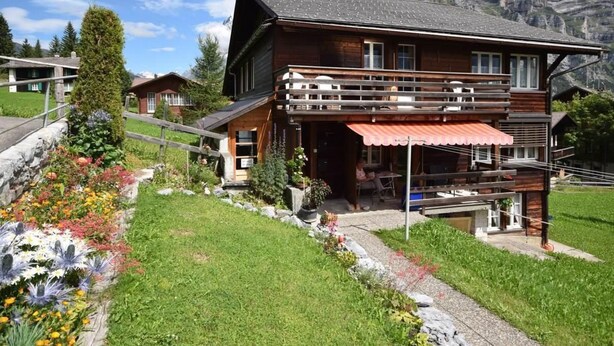 Un chalet in legno con un balcone, immerso in un giardino fiorito, con montagne sullo sfondo.