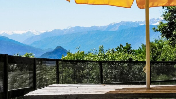 Een houten terras met een gele parasol, een zwart hek en uitzicht op een berg.