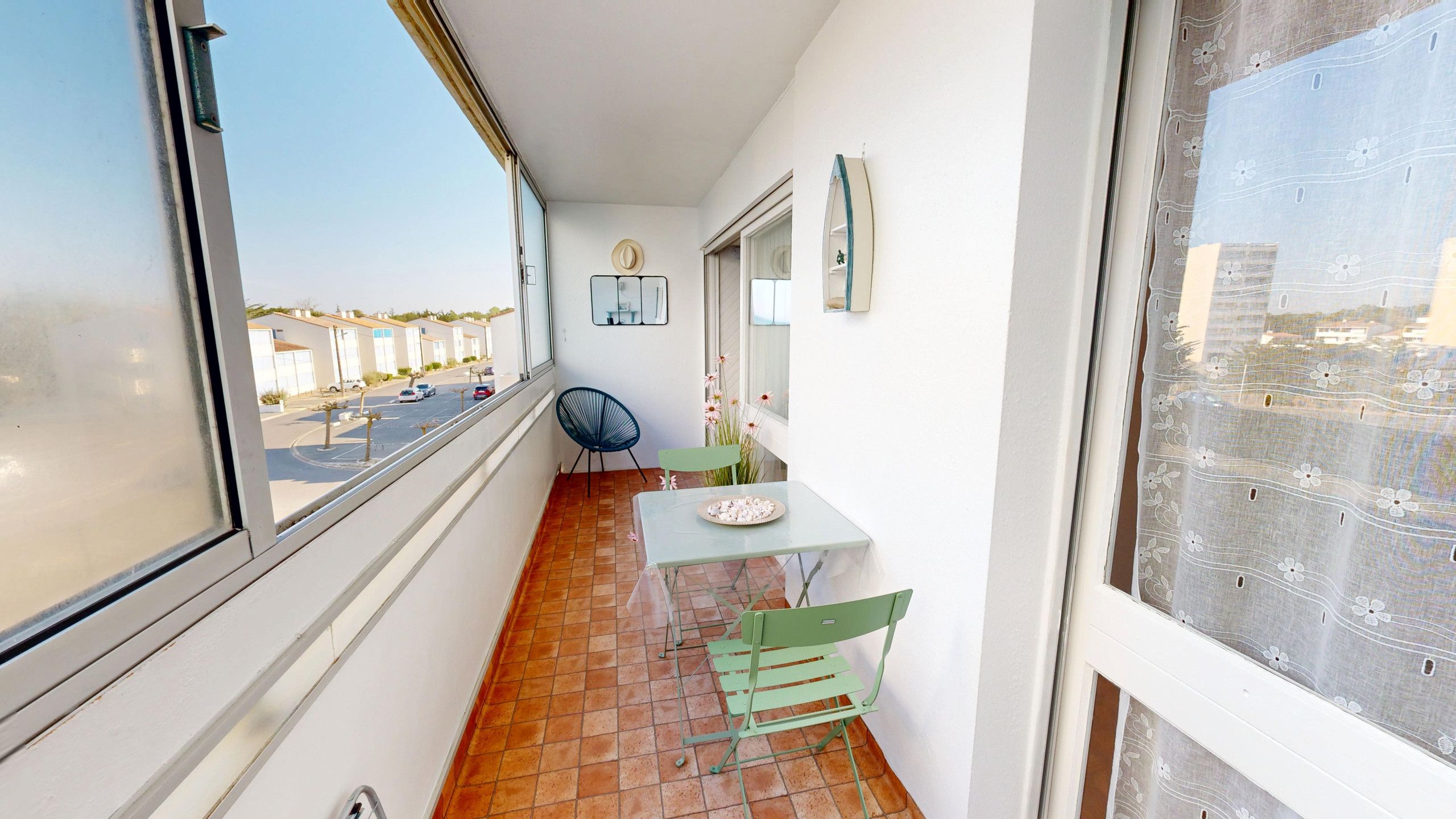 A narrow balcony with a table and chairs, a view of the street, and a small garden area.