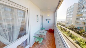 A balcony with a table and chairs, a window with sheer curtains, a small shelf, and a view of a building and greenery. - Air Marin - Apartment Balcony Ocean View (Saint-Hilaire-de-Riez)