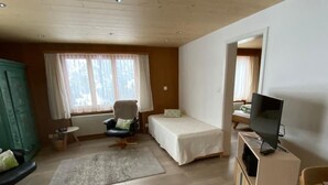 Una habitación pequeña y luminosa con una cama, una silla, un televisor y un escritorio.