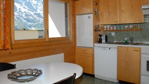 Een kleine keuken met houten kastjes, een witte koelkast, een koffiezetapparaat en een eettafel met stoelen.