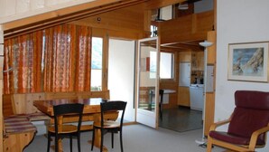 Een knus vertrek met een houten eettafel en stoelen, een rode fauteuil, en zicht op een keuken en eetruimte door een open deur.