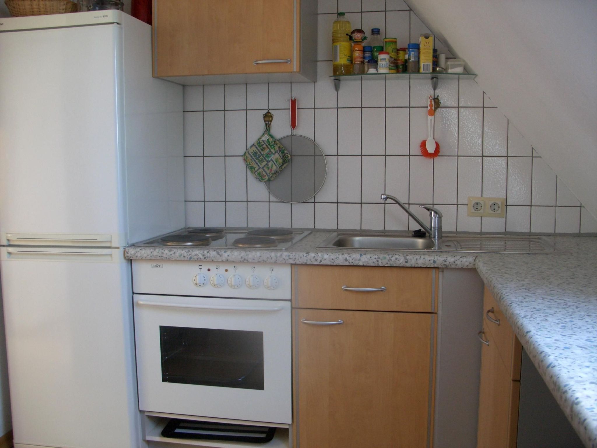 A kitchen with a white refrigerator, stove, oven, and wooden cabinets. There is a sink with a faucet, and a shelf above the sink holding various kitchen items.