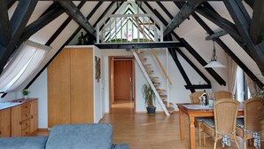 Een zolderkamer met zichtbare houten balken, een trap, een zithoek met rieten stoelen en een grijze bank.
