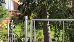 Een houten terras met een metalen hekwerk, een bloempot en uitzicht op een achtertuin met gras en bomen.