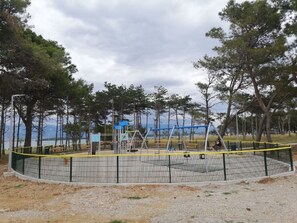 Ein von einer Hecke umgebener Spielplatz mit Schaukeln und einer blauen Konstruktion, umgeben von Bäumen und einem Gewässer.