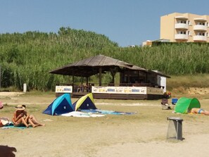 Ein Strand mit einem hölzernen Pavillon, Zelten und einem Gebäude im Hintergrund.