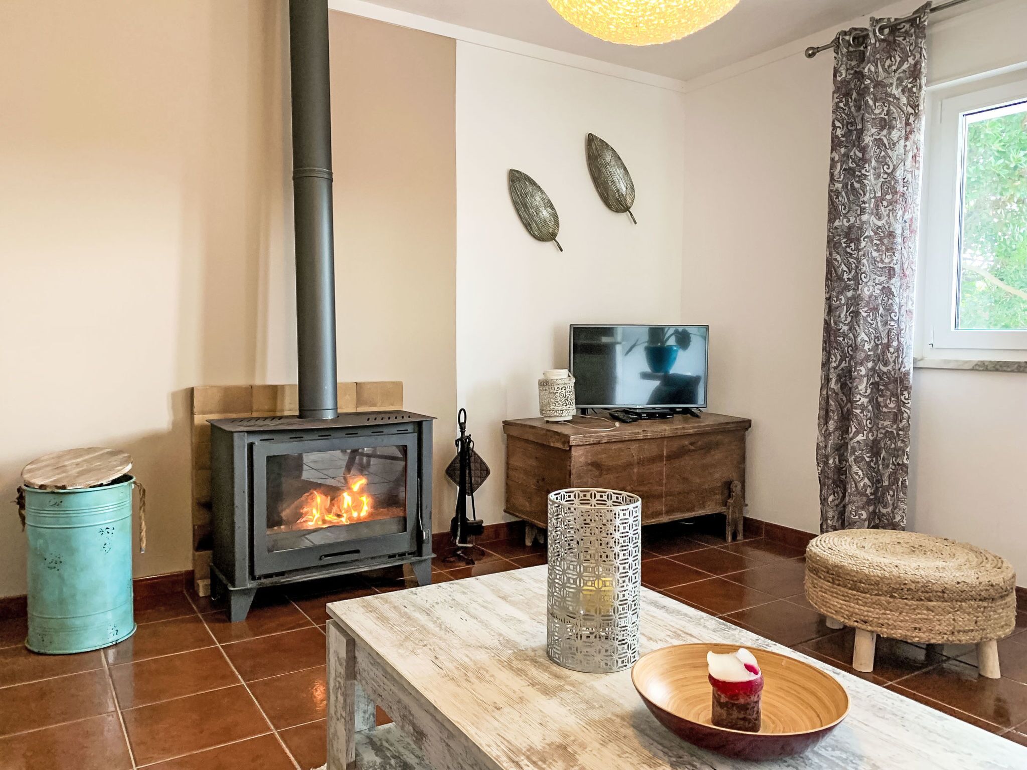 A living room with a wood-burning stove, a television, a table with a bowl, and a window with curtains.