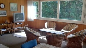 Une pièce confortable avec une télévision, des fauteuils en osier, une table et de grandes fenêtres donnant sur la forêt.