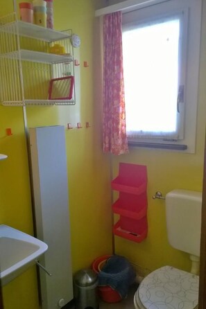 Ein kleines Badezimmer mit gelber Wand, Waschbecken in Weiß, Toilette und Fenster mit rosa Vorhang. Ein Ablagebereich bietet Platz für Toilettenartikel.