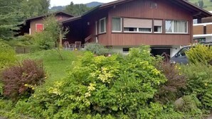 Une maison à deux étages avec un jardin devant, une voiture garée dans l’allée et un bâtiment rouge en arrière-plan.
