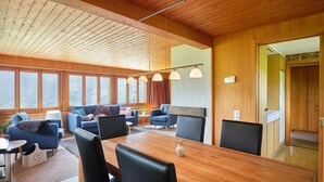 Ein geräumiges Wohnzimmer mit Holzboden, großen Fenstern und einem Essbereich mit Holztisch und -stühlen.