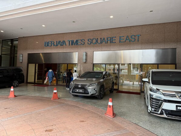 Berjaya Times Square East -rakennuksen ulkopuoli: moderni julkisivu, suuri lasinen sisäänkäynti ja kaksi pysäköityä autoa.