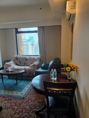 A hotel room with a sofa, a dining table with chairs, a small rug, and a window with a view of buildings. - Homestay Suite At Times Square KL (Kuala Lumpur)