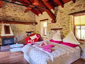 Une chambre avec un mur en pierre, une cheminée, un lit avec une literie rose et blanche, une chaise rouge et une petite table ronde.