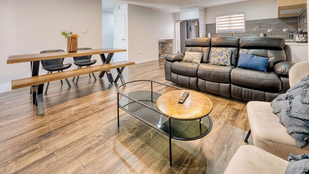 A modern living room with a leather sofa, a glass coffee table, a dining table with chairs, and a kitchen area in the background. - Boutique Hillside Heaven near AKLD Airport (Papatoetoe Auckland)