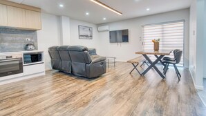 A modern kitchen with built-in appliances, a dining area with a table and chairs, a reclining chair, and a large window with blinds. - Boutique Hillside Heaven near AKLD Airport (Papatoetoe Auckland)