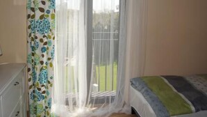 Ein Schlafzimmer mit Bett, einem Fenster mit durchsichtigen Vorhängen und Blick auf eine grüne Außenanlage.