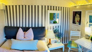 Una camera da letto con una parete a righe, un letto con molti cuscini, un comodino con una lampada, un tavolino con un vaso di fiori e una sedia.