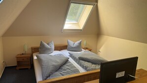 Una camera da letto piccola e accogliente con soffitto a falde, una finestra a lucernario, un letto con biancheria grigia, un comodino con una lampada e un televisore su un supporto in legno.