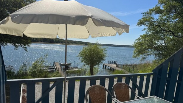 Un patio con tavolo e sedie in vetro, un ombrellone e vista su un lago con barche e un molo.