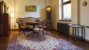 Una habitación acogedora con un sofá tradicional, una mesa de comedor y sillas. Hay un ventanal con vistas, un radiador y un reloj de pared.