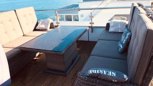 A sunlit outdoor seating area with a view of the sea, featuring a sofa, a table, and a wicker basket. - Schwimmendes Haus Elandi by Interhome (Bitterfeld)