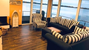 A cozy living room with a black sofa and striped armchairs, large windows offering a view of a lake, and a wooden floor. - Schwimmendes Haus Elandi by Interhome (Bitterfeld)