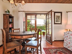 Uma sala de jantar com mesa e cadeiras de madeira, uma estante de livros, um lustre e vista através de uma porta aberta.