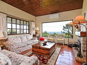 Uma sala de estar acolhedora com sofá, mesa de centro de madeira, cadeiras de vime, lareira e uma janela ampla com vista.