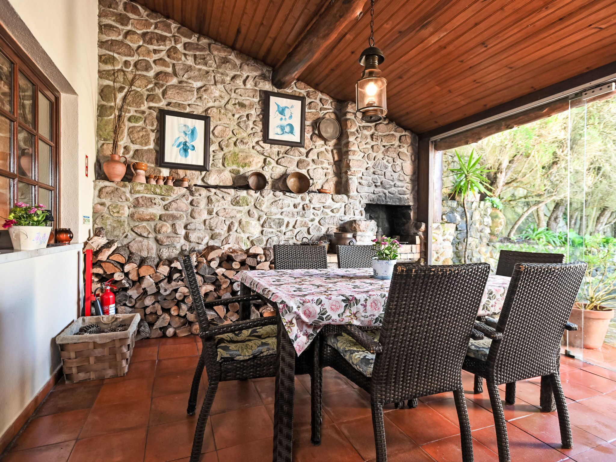 A stone wall, wooden ceiling, wicker chairs, a table with a floral tablecloth, and a hanging light fixture.