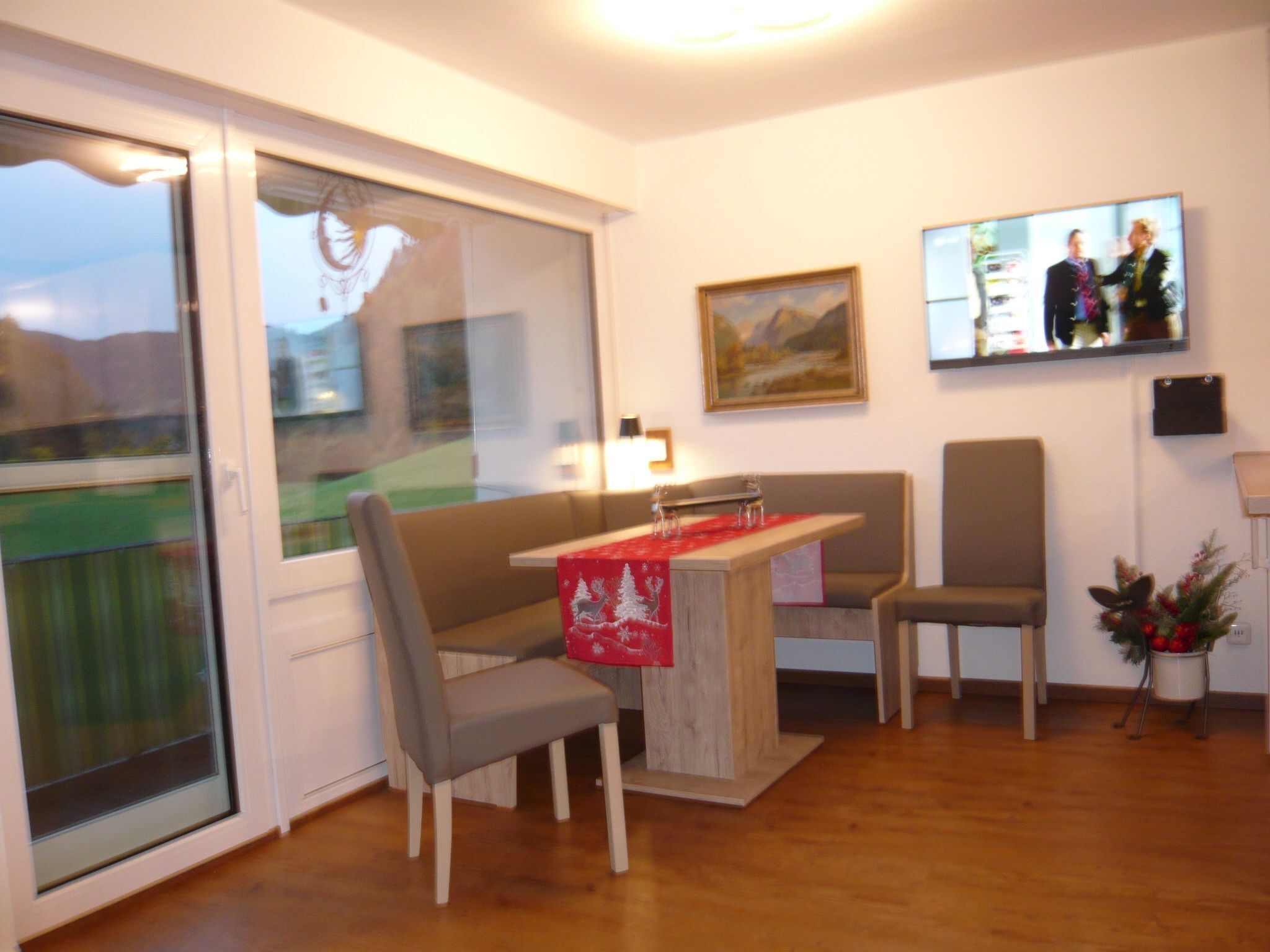 Un salon moderne avec une table à manger en bois, deux chaises grises, un chemin de table rouge, une grande fenêtre donnant sur un paysage verdoyant, un tableau accroché au mur et un téléviseur fixé au-dessus de la table.
